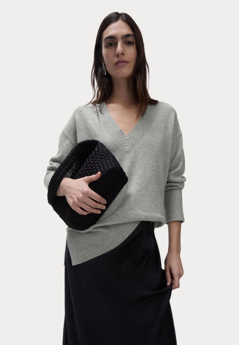 Lichtgrijze, oversized trui met een V-hals en geribbelde details, gecombineerd met een zwarte gehaakte clutch. De outfit bevat een zwarte rok.