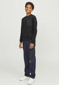 Svart sweatshirt med framficka, kombinerad med mörkblå jeans. Modellen bär vita sneakers och står framför en neutral bakgrund.