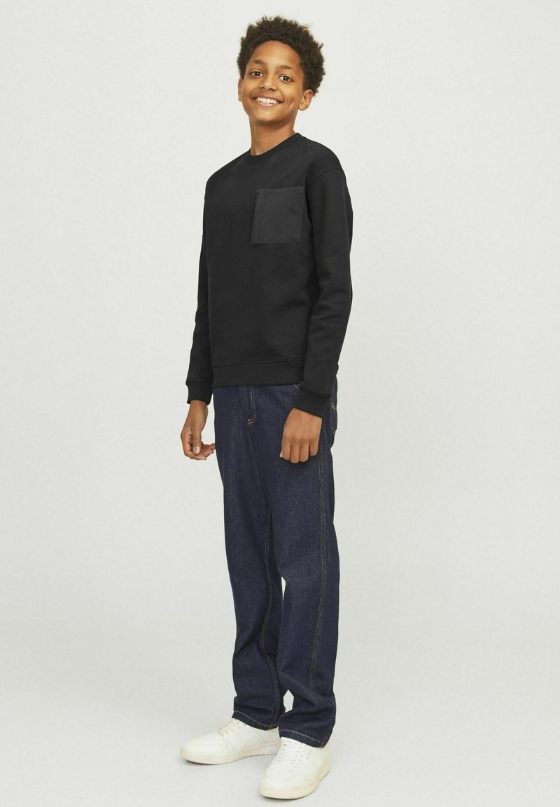 Svart sweatshirt med framficka, kombinerad med mörkblå jeans. Modellen bär vita sneakers och står framför en neutral bakgrund.