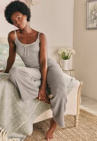 Grauer, gerippter Jumpsuit mit dünnen Trägern und einem Frontknotendetail. Hat einen lockeren Schnitt und weite Beine. Auf einem Bett mit einer weichen Decke drapiert.