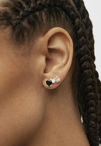 Orecchini a bottone in argento che presentano un cuore nero, una perla bianca e accenti in pietra trasparente, con i capelli intrecciati e una carnagione chiara.