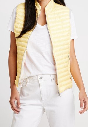 Une femme porte un gilet matelassé sans manches jaune pâle par-dessus un T-shirt blanc et un jean blanc.