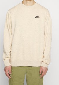 Sweatshirt crème avec une texture mouchetée, manches longues et poignets en côtes. Présente un petit logo Nike noir sur la poitrine.