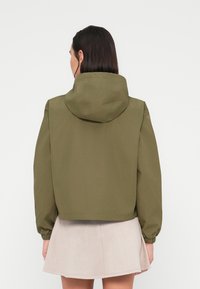 Γυναίκα που φοράει μπουφάν με κουκούλα χακί με ελαστικά μανίκια και φούστα ανοιχτού μπεζ χρώματος, εμφανιζόμενη από πίσω με φόντο λευκό.