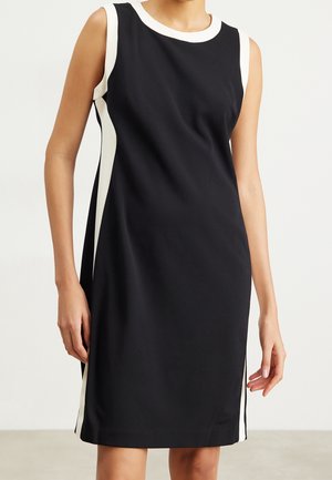 Czarna sukienka bez rękawów, o długości do kolan, z białym wykończeniem na dekolcie, otworach na ręce i bocznych panelach, noszona przez stojącą osobę.