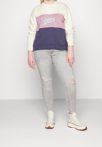 Sudadera morada, crema y azul marino con el logo de "Adidas"; puños y dobladillo acanalados. Combinada con jeans pitillo grises claros rasgados y zapatillas blancas.