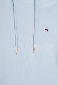 Sudadera azul claro hecha de un tejido suave, con un bolsillo frontal y drawstring. Presenta un pequeño logo bordado con acentos en rojo, blanco y azul marino.