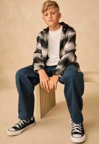 Chemise en flanelle à carreaux noir et blanc superposée sur un T-shirt blanc, assortie à un pantalon bleu foncé et des baskets noires. Assis sur une surface beige.