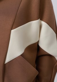 Gros plan sur un tissu marron avec une large bande diagonale blanche et un détail de poche cousue en bas à gauche.