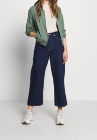 Zelená bomber bunda, světlé tričko a tmavé široké džíny. Bílé tenisky doplňují outfit. Hladká textura na oblečení.