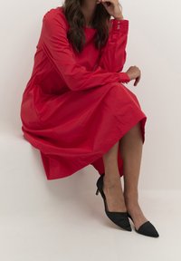 Rode jurk met lange mouwen en een relaxte pasvorm, voorzien van knoopmanchetten en een knielange zoom, gecombineerd met zwarte hoge puntige hakken.