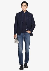 Marineblå tekstureret blazer, mørk sweater, blå jeans og sorte sko; holder en foldet avis; tætsiddende design med moderne silhuet.