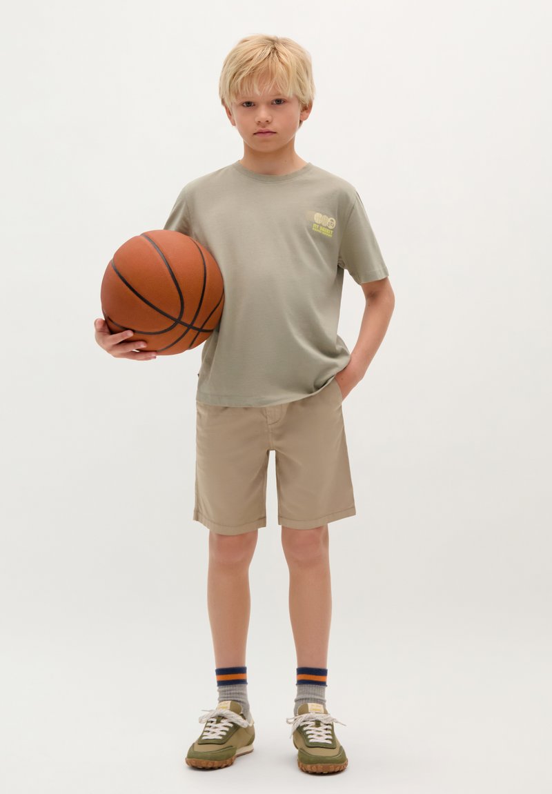 Helles grünes T-Shirt mit kurzen Ärmeln, beige Shorts, Basketball in einer Hand, grün-weiße Sneakers und graue Socken mit blauen und orangen Streifen.