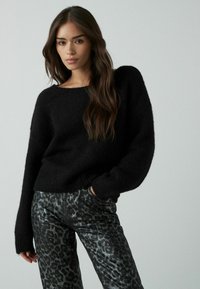 Zwarte gebreide trui met een brede halslijn en losse mouwen, gecombineerd met zwarte leren broek met een metallic luipaardprint patroon.