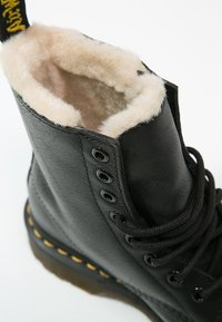 Botte en cuir noir avec doublure en fausse fourrure, bout rond, fermeture à lacets, semelle cousue jaune, œillets métalliques et étiquette de marque jaune sur le talon.