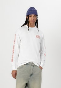 Long-sleeve white t-shirt with red graphic text on sleeves and chest; paired with faded blue jeans; model wearing a blue striped beanie.