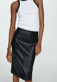 Femme portant un haut blanc sans manches avec une bordure noire et une jupe crayon en cuir noir taille haute sur un fond uni.