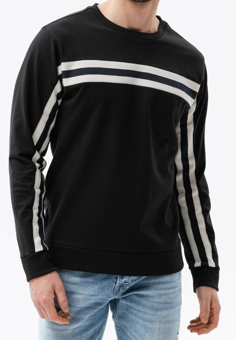 Homme portant un pull noir à manches longues avec des rayures blanches et bleu foncé sur la poitrine et le long des manches, associé à un jean bleu clair.