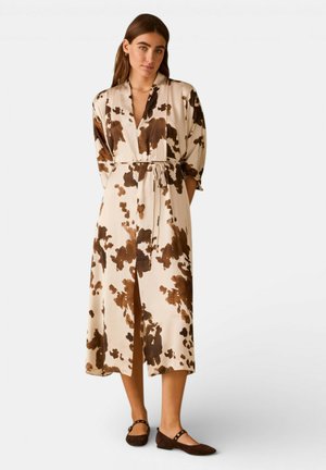 Donna in piedi che indossa un vestito midi beige con macchie astratte marroni, maniche lunghe, scollo a V, cintura in vita e scarpe basse marrone scuro.