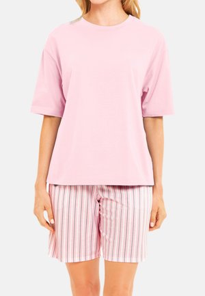 Frau trägt ein hellrosa kurzärmeliges T-Shirt und rosa, weiß-schwarze, vertikal gestreifte Shorts vor einem einfarbigen Hintergrund.