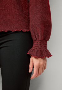 Rode, glanzende lange mouwen top met smockmanchetten en geschulpte zoom. Gecombineerd met zwarte, gestructureerde broeken. Close-up van de mouw en hand.