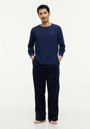 Mand stående barfodet iført en marineblå langærmet top og matchende marineblå pyjamasbukser med små flagmønstre. Hænderne i lommerne.
