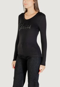 Maglietta nera a maniche lunghe con scollo rotondo, presenta il logo "Guess" impreziosito da strass, design aderente, abbinata a jeans scuri.