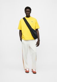 Νέος άνδρας που φοράει κίτρινη μπλούζα Adidas, λευκό παντελόνι φόρμας με ρίγες, κόκκινα αθλητικά παπούτσια και μαύρη τσάντα ώμου μπροστά σε λευκό φόντο.