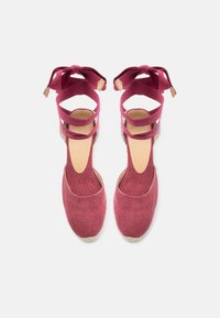Paire d'espadrilles rouges en toile avec liens à nouer autour de la cheville et talons compensés tissés, vues de dessus sur un fond blanc.