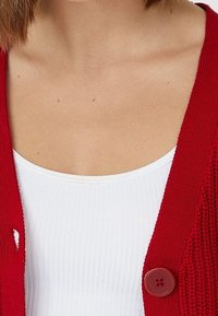 Cardigan rouge en tricot avec une fermeture à boutons bien visible, porté au-dessus d'un débardeur blanc à côtes. Le tissu a un motif texturé.
