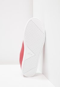 Röd sneaker med en vit texturerad sula som har en krokodillogo, placerad upprätt mot vita väggar.