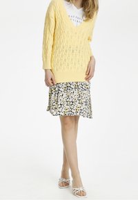 Gelber Strickpullover mit einem Rautenmuster, kombiniert mit einem schwarzen Blumenrock, der gelbe und weiße Blumen zeigt. Weiße Sandalen mit Riemchen runden das Outfit ab.