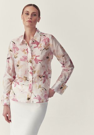 Kobieta z zaczesanymi do tyłu włosami, ubrana w różowo-beżową bluzkę w kwiaty z długim rękawem oraz białą spódnicę, stoi z jedną ręką na biodrze.