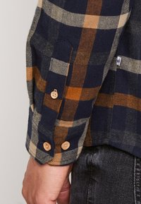 Camicia di flanella a quadri blu con motivi marroni e beige, polsino con bottoni, un sottile motivo a spina di pesce e texture morbida.