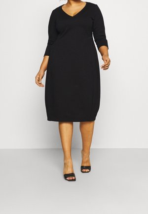 Czarna sukienka z dekoltem w serek, rękawami do połowy łokcia oraz dopasowanym krojem. Gładka tkanina z detalami na bocznych szwach, zestawiona z czarnymi sandałkami na obcasie z odkrytymi palcami.