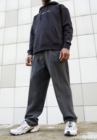 Hombre con una sudadera negra, pantalones grises y zapatillas blancas de pie sobre una superficie de grava frente a un fondo de pared de azulejos.