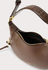 Geantă de mână din piele maro cu o formă curbată, fermoar auriu și căptușeală interioară. Marcajul este afișat în partea de jos, inclusiv o curea reglabilă.