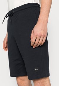 Tmavě navy bavlněné šortky mají elastický pas s tkaničkou a malou nášivku na spodní straně. Design je jednoduchý a sportovní.
