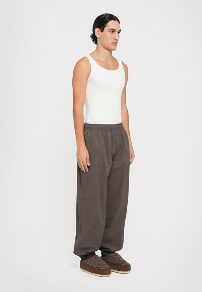 Hvid tanktop, løse brune bukser med elastisk talje, detaljeret broderi og brune tøfler med mønstrede accenter.