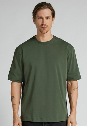 Mężczyzna o jasnobrązowych włosach, ubrany w jednolitą ciemnozieloną koszulkę z krótkim rękawem, stojący na jasnoszarym tle.