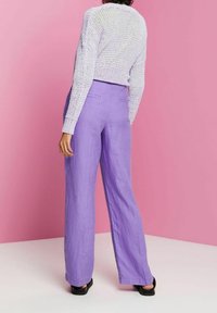 Osoba ubrana w biały sweter z dzianiny i wysokie, szerokie lawendowe spodnie, stojąca na różowym tle.