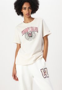 Kremfarget t-skjorte med korte ermer, med et sentralt trykk av en tiger og "TOMMY JEANS" i rødt tekst. Mykt stoff, avslappet passform.