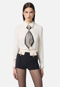 Blusa crema con una cravatta a righe bianche e nere, abbinata a pantaloni corti blu navy a vita alta con dettagli crema e accessori in oro.