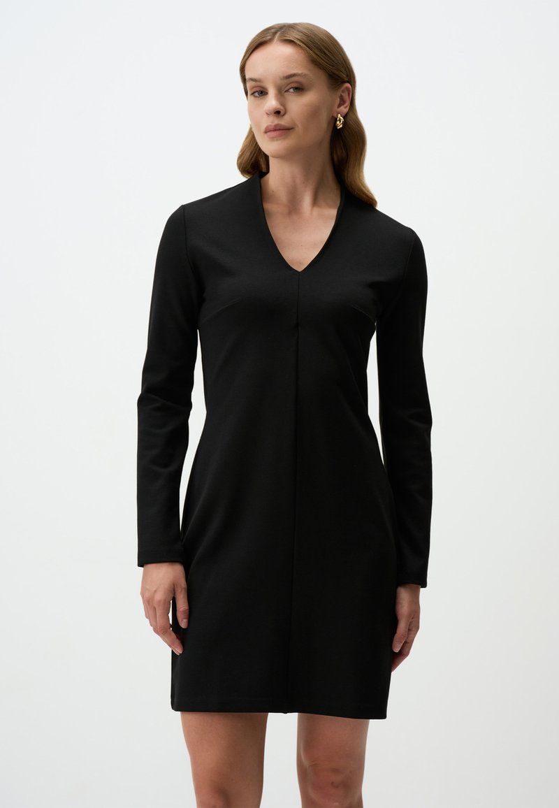 Robe noire à manches longues avec un décolleté en V, en tissu lisse. Elle présente une silhouette ajustée et une couture centrale sur le devant.