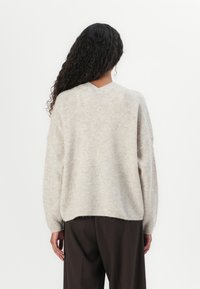 Knit sweater in light beige, featuring a V-neck, loose fit, ribbed texture, and dropped shoulders. Paired with dark brown trousers.