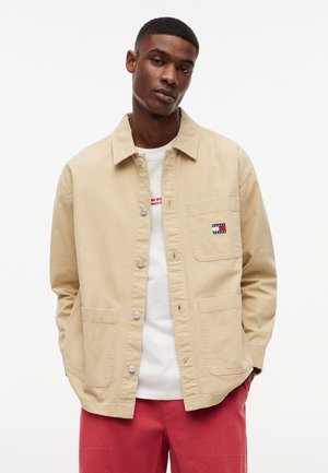 Junger Mann trägt eine beige Jacke mit Knöpfen über einem weißen T-Shirt und roten Hosen, steht vor einem schlichten weißen Hintergrund.