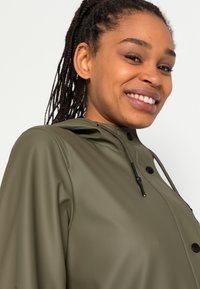 Imperméable vert olive avec capuche, présentant des accents noirs, un cordon de serrage à bascule et une texture lisse. Gros plan sur le col et la zone des épaules.