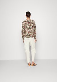 Langärmlige Bluse mit Blumenmuster auf cremefarbigem Hintergrund, die mit mehrfarbigen Blumenmotiven verziert ist. Kombiniert mit locker sitzenden weißen Hosen und braunen Absatzsandalen.