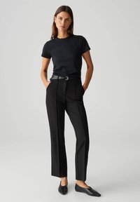 Zwarte, getailleerde t-shirt gecombineerd met zwarte, op maat gemaakte broek met zijplooien en een zwarte riem. Het model draagt platte zwarte schoenen. Neutrale achtergrond.