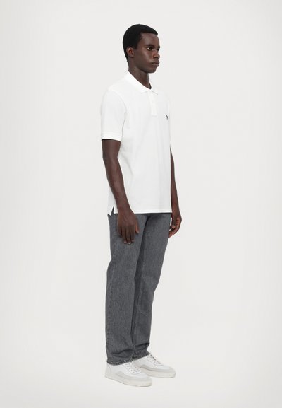 Valge polo särk kraega, hallide denim teksade ja valgete tossudega, seismas lihtsa heleda tausta vastas.